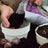 Hands filling a white basket-weave planter with potting soil from a purple bag, with several purple flower bulbs visible in the foreground and additional planters nearby